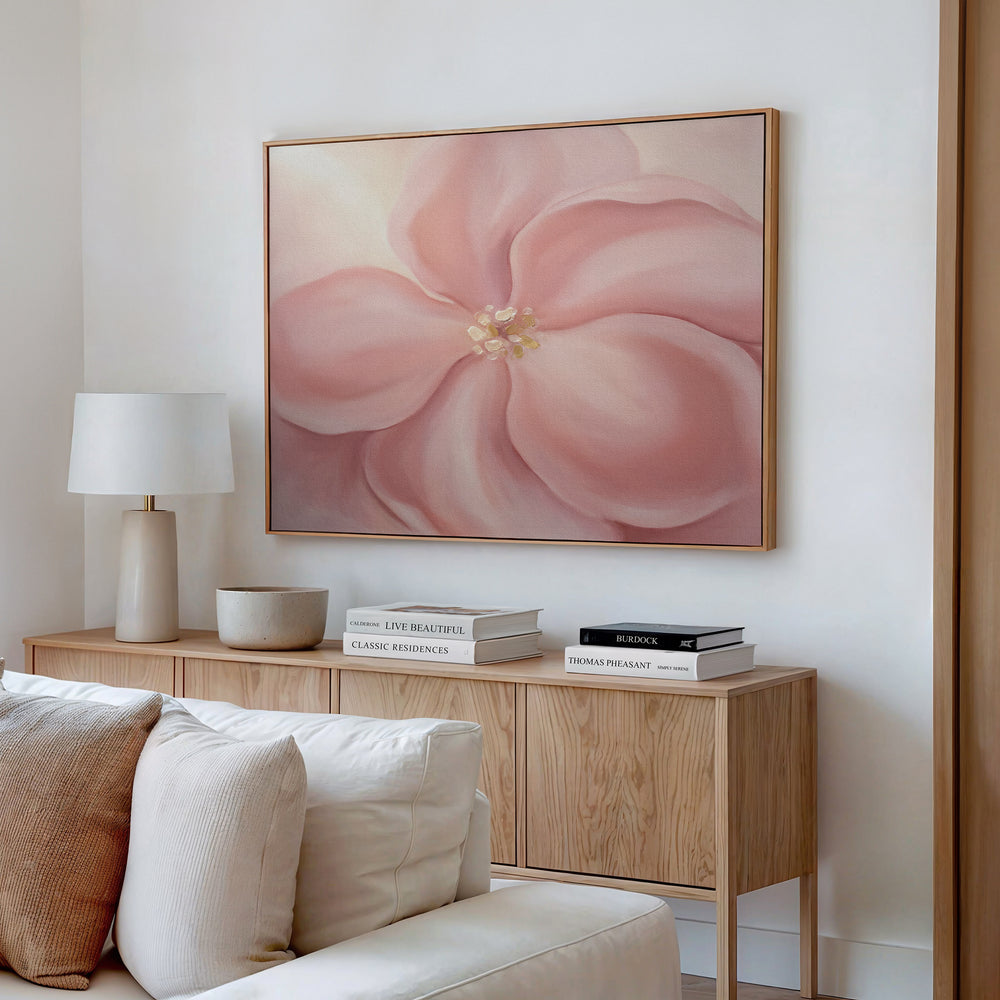 A cozy living room corner features a light wood sideboard with stacked books, a white lamp, and a bowl, set beneath the Blushing Magnolia Canvas Art - Horizontal, adding soft romantic wall art to the tranquil white space.
