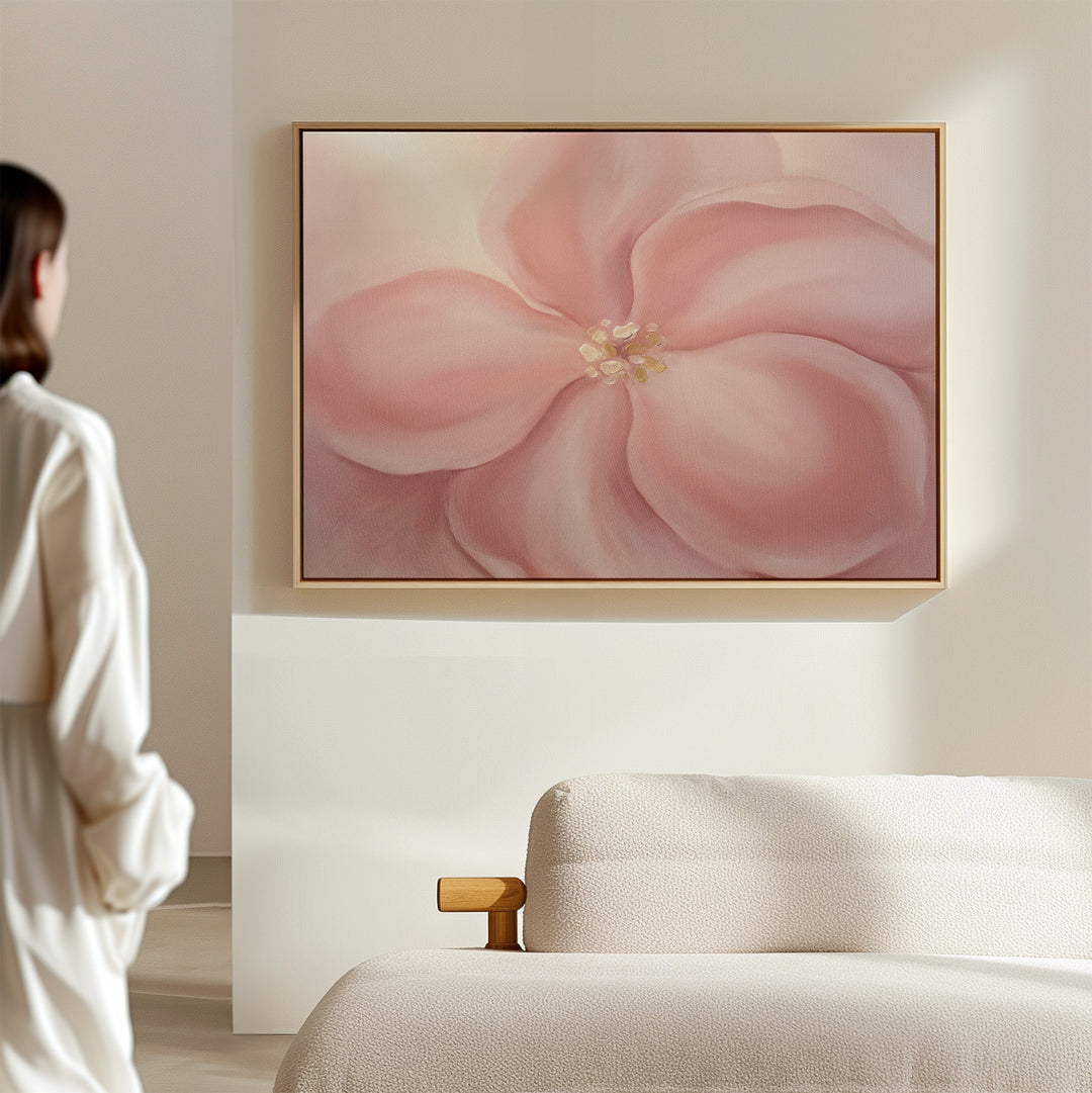 A person in a white robe stands in a softly lit room, admiring the Blushing Magnolia Canvas Art - Horizontal hanging above a beige sofa.