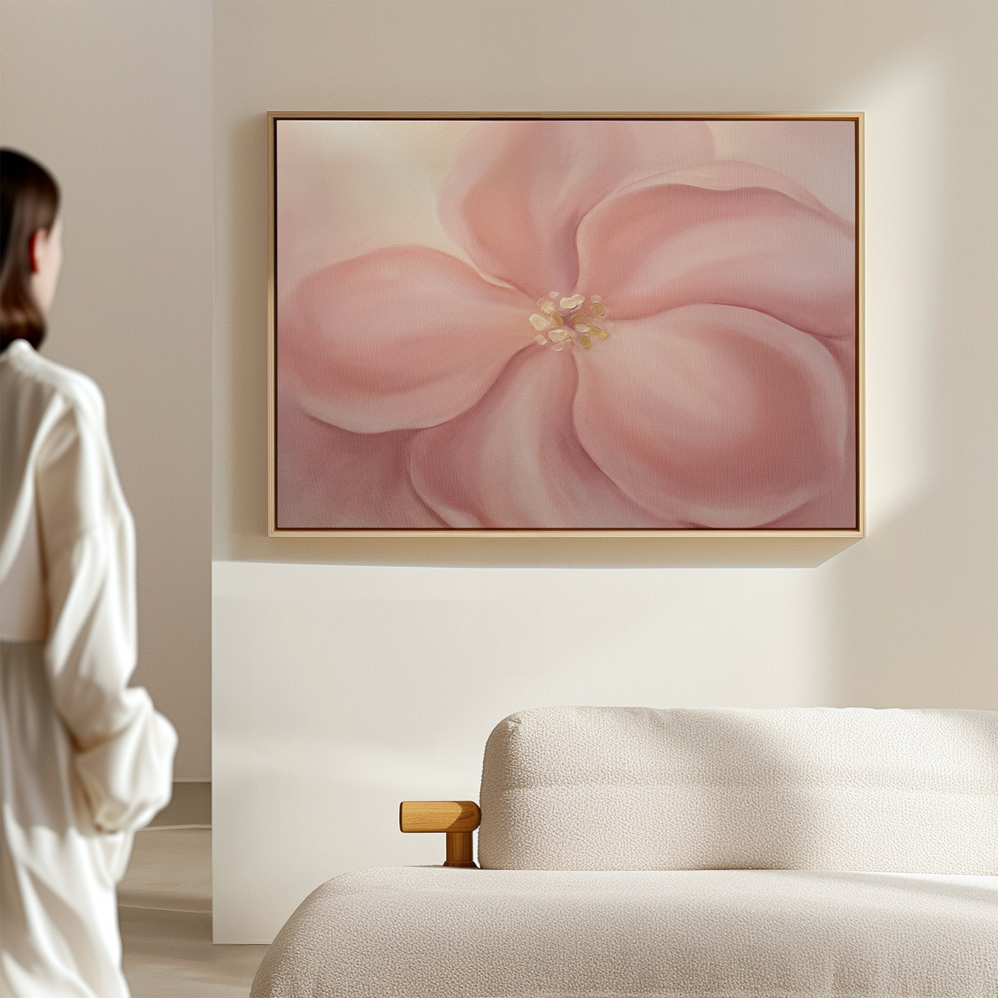 A person in a white robe stands in a softly lit room, admiring the Blushing Magnolia Canvas Art - Horizontal hanging above a beige sofa.