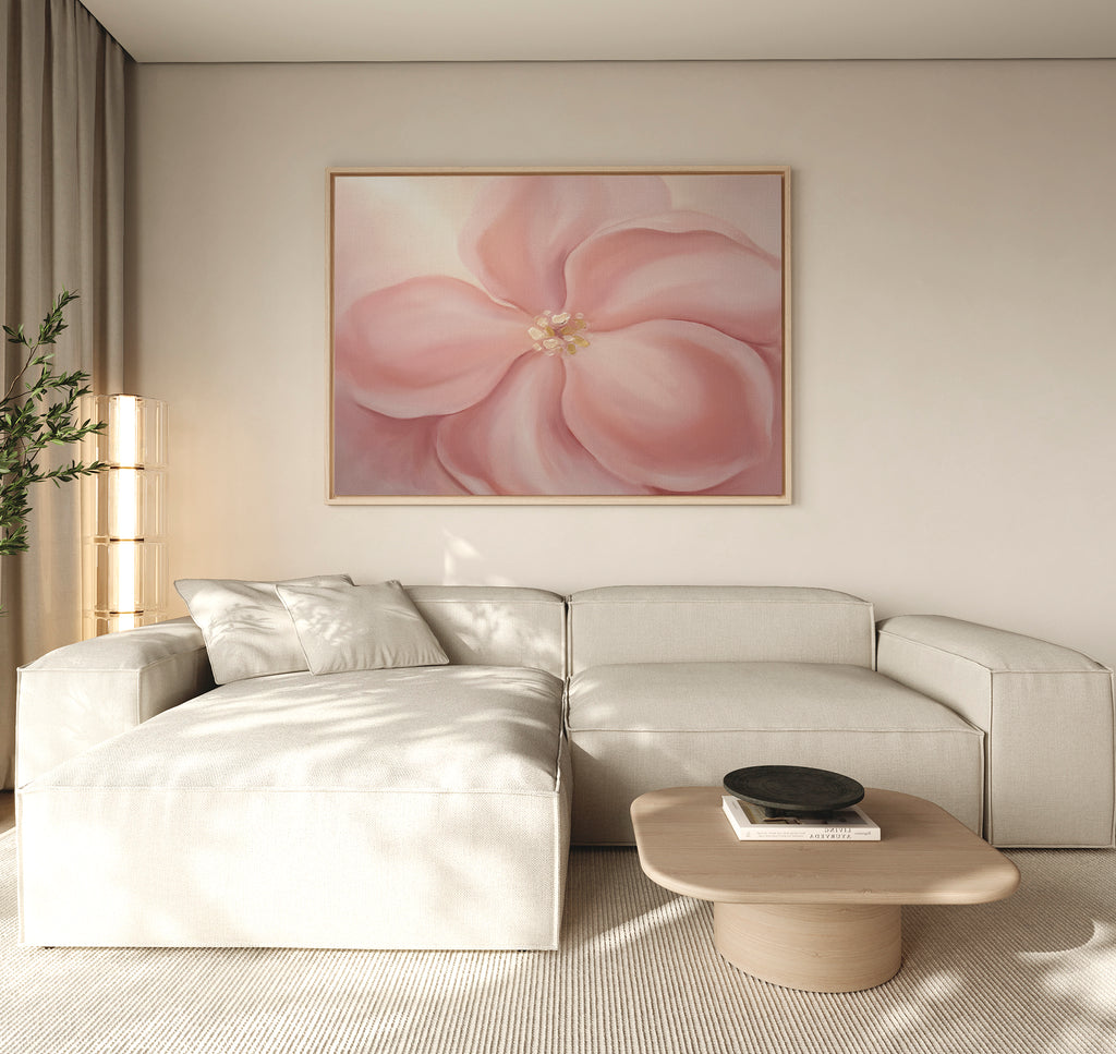 A modern living room features a beige sectional, a light wood coffee table, and the Blushing Magnolia Canvas Art - Horizontal on the wall. Soft neutrals and natural light enhance the calm, minimalist vibe.