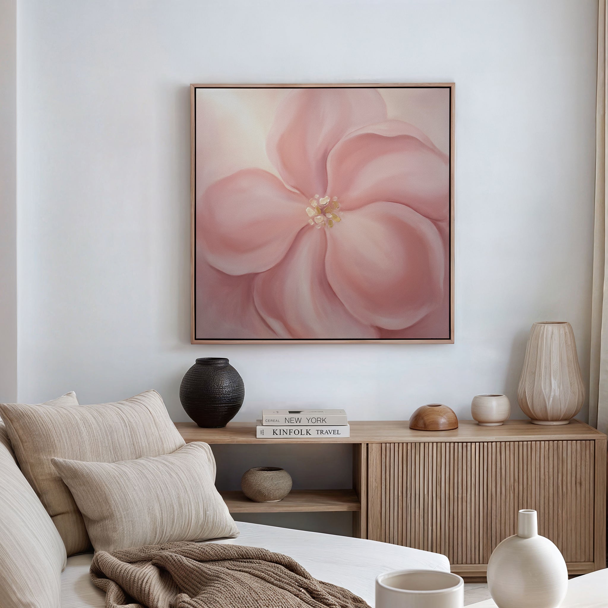 A cozy living room featuring a beige sofa, neutral cushions, wooden console table, decorative vases, stacked books, and the Blushing Magnolia Canvas Art - Square as soft romantic wall art above the space.