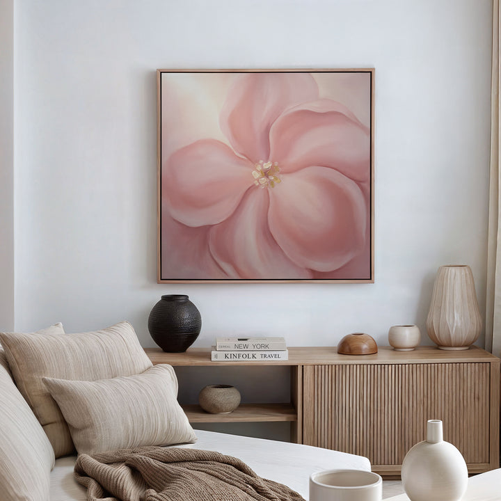 A cozy living room featuring a beige sofa, neutral cushions, wooden console table, decorative vases, stacked books, and the Blushing Magnolia Canvas Art - Square as soft romantic wall art above the space.