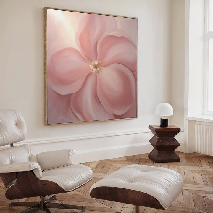 A modern, minimalist room featuring a white lounge chair and ottoman, wooden side table with a small white lamp, and the Blushing Magnolia Canvas Art - Square as soft romantic wall decor. Light wood floors and a tall window complete the look.