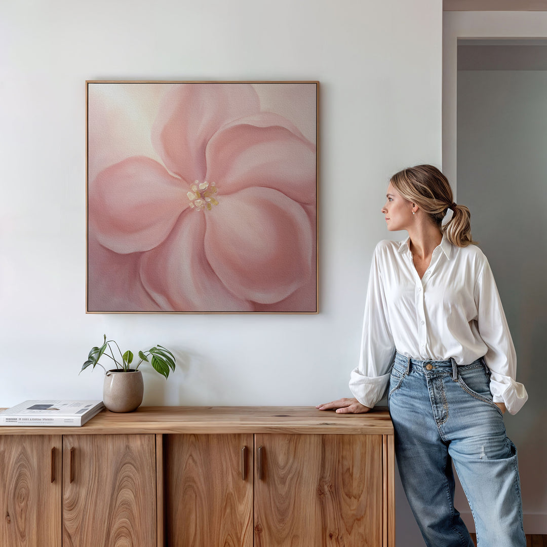 A woman in a white blouse and jeans stands by a wooden cabinet, admiring the Blushing Magnolia Canvas Art - Square on the wall. A potted plant and book sit atop the cabinet in this bright, minimalist space.