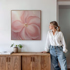 A woman in a white blouse and jeans stands by a wooden cabinet, admiring the Blushing Magnolia Canvas Art - Square on the wall. A potted plant and book sit atop the cabinet in this bright, minimalist space.