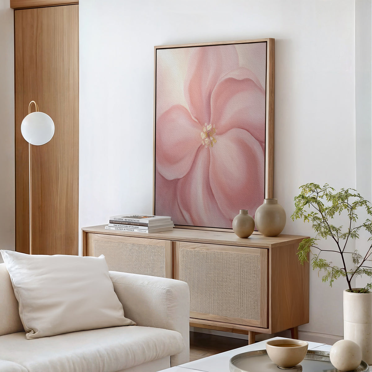 A modern living room with light wood furniture, a white sofa with cream cushion, a sideboard displaying vases and books, soft romantic wall art featuring the Blushing Magnolia Canvas Art - Vertical above, and a small green plant in the corner.