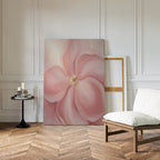 The Blushing Magnolia Canvas Art - Vertical, with its soft pink flower, leans against a white paneled wall next to an empty frame. A cushioned chair and small black table with candles and books sit nearby on a wooden herringbone floor.