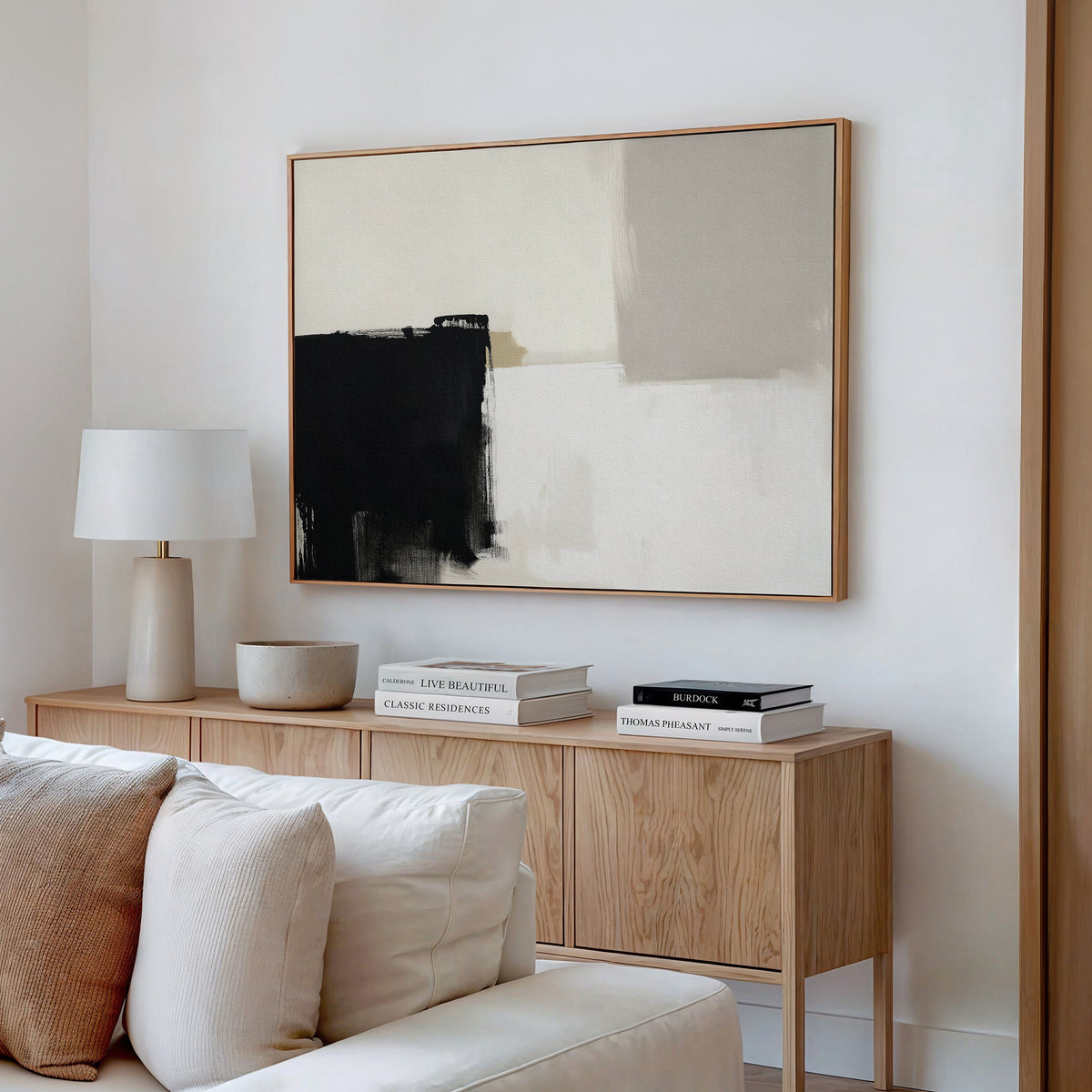 Minimalist living room with a light wood sideboard, white table lamp, decorative bowl, stacked books, and the Bold Division Canvas Art - Horizontal featuring black, beige, and cream on a white wall. White sofa cushions add comfort—ideal for modern interiors.