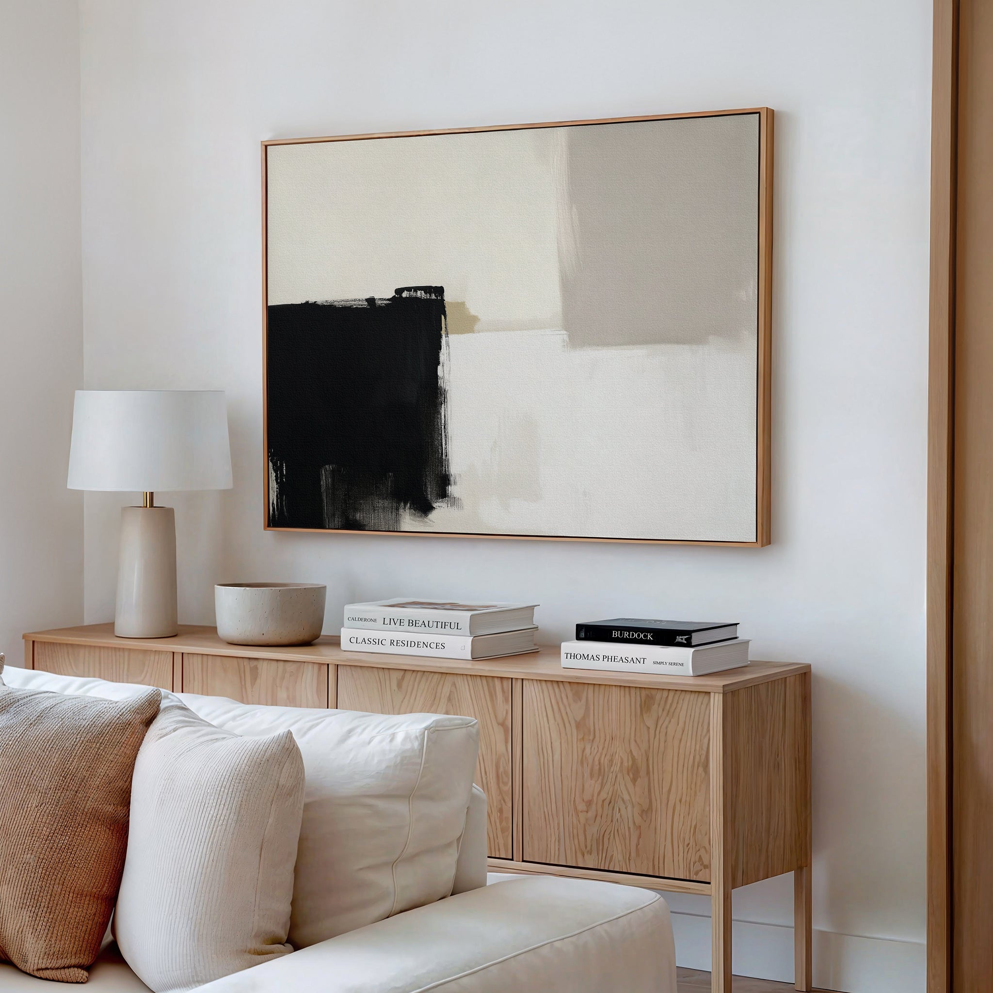 Minimalist living room with a light wood sideboard, white table lamp, decorative bowl, stacked books, and the Bold Division Canvas Art - Horizontal featuring black, beige, and cream on a white wall. White sofa cushions add comfort—ideal for modern interiors.