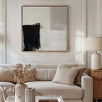 A modern living room featuring a beige sofa, neutral pillows, a wooden side table with a ceramic lamp, dried flowers in a vase, and the Bold Division Canvas Art - Square on the wall elevates contemporary style.