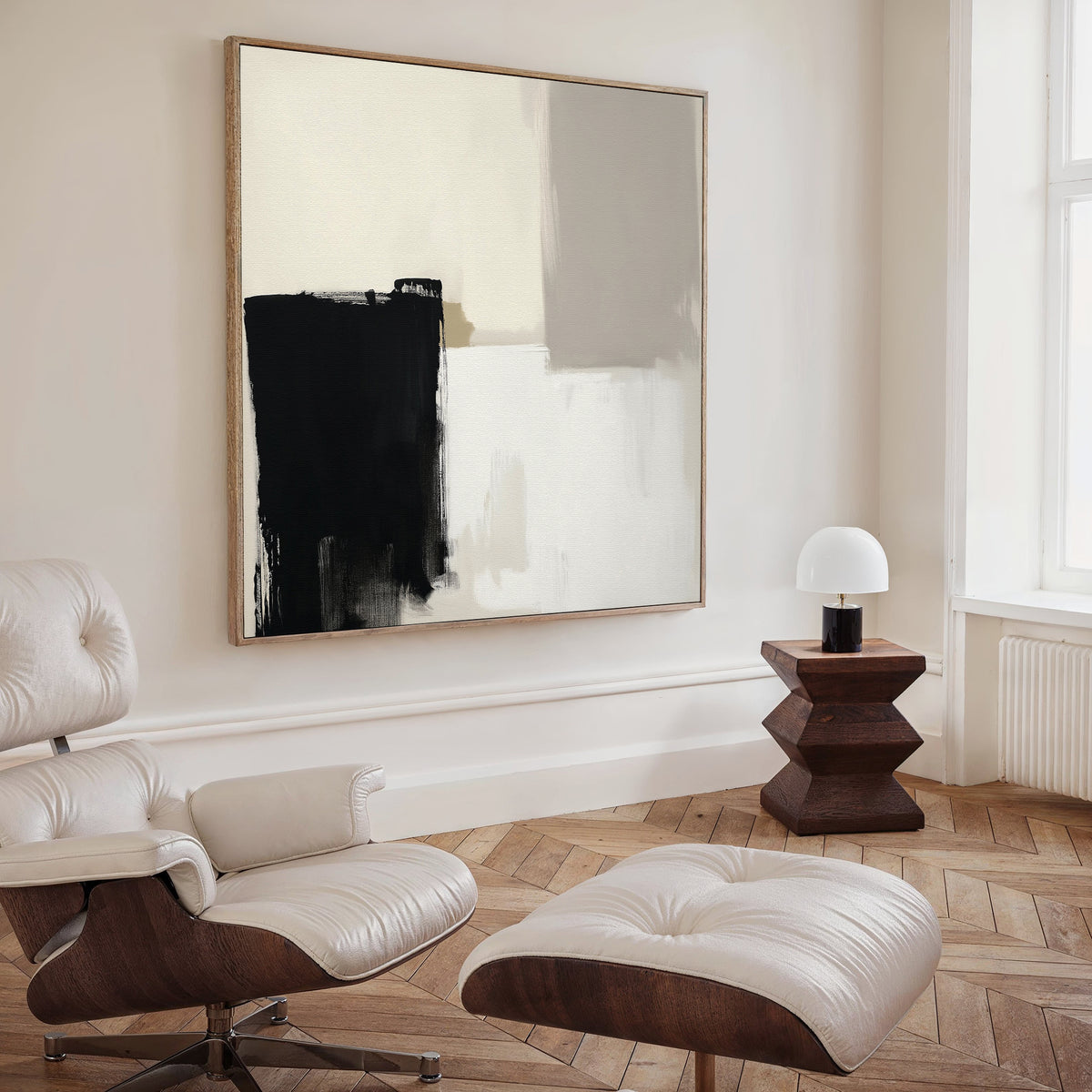 A modern living room with a white leather lounge chair and ottoman, wooden floors, a geometric side table with a white lamp, and Bold Division Canvas Art - Square—a striking black, white, and beige contemporary print—highlighting modern decor.
