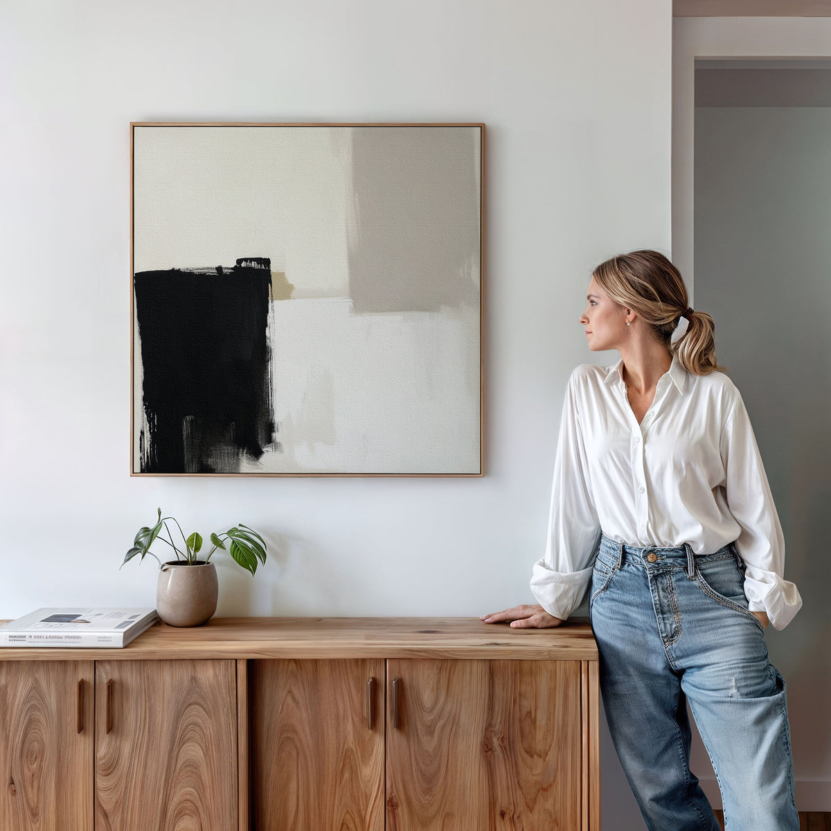 A woman in a white blouse and jeans leans against a wooden cabinet, admiring the Bold Division Canvas Art - Square on the wall. A plant and book on the cabinet enhance this minimal, modern living room's style.