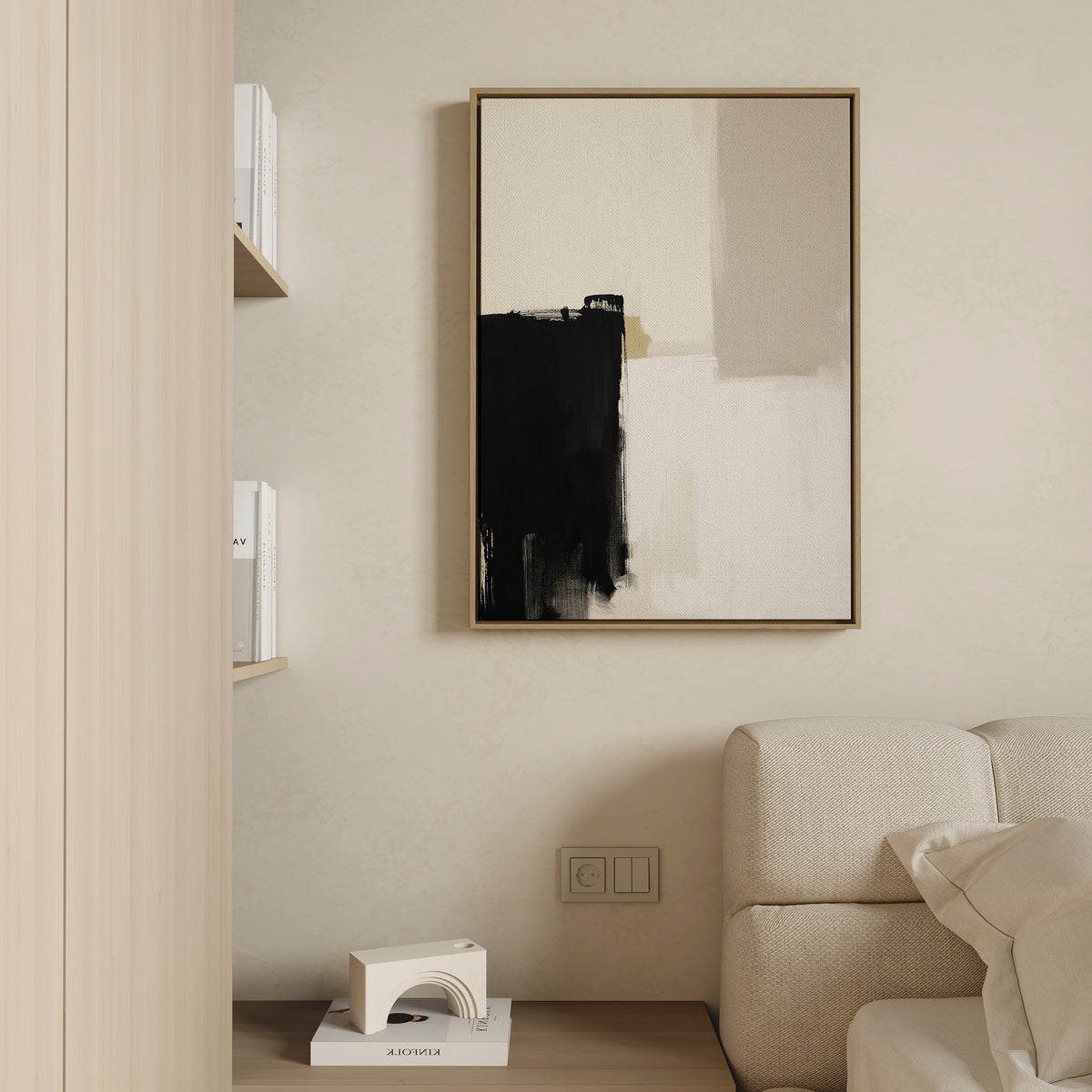 A minimalist interior features a beige couch, a light wood shelf with books and a sculpture, and the Bold Division Canvas Art - Vertical in black, white, and beige on a cream wall above electrical outlets.