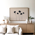 A modern living room with a light wood sideboard, neutral vases, a lamp, a plant, and the Botanic Abstract Canvas Art—a black minimalist floral print—framed above, paired with a white sofa and cream cushions in front.