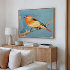 A modern living room featuring a wooden sideboard, books, lamp, bowl, and the Bright Songbird Canvas Art - Horizontal—a large colorful bird on a branch against blue—adds charm to the space.