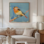 A modern living room with a beige sofa and neutral pillows features the Bright Songbird Canvas Art - Square, a colorful painting of a bird on a branch against blue, hung above the sofa. Dried flowers and a table lamp complete the decor.