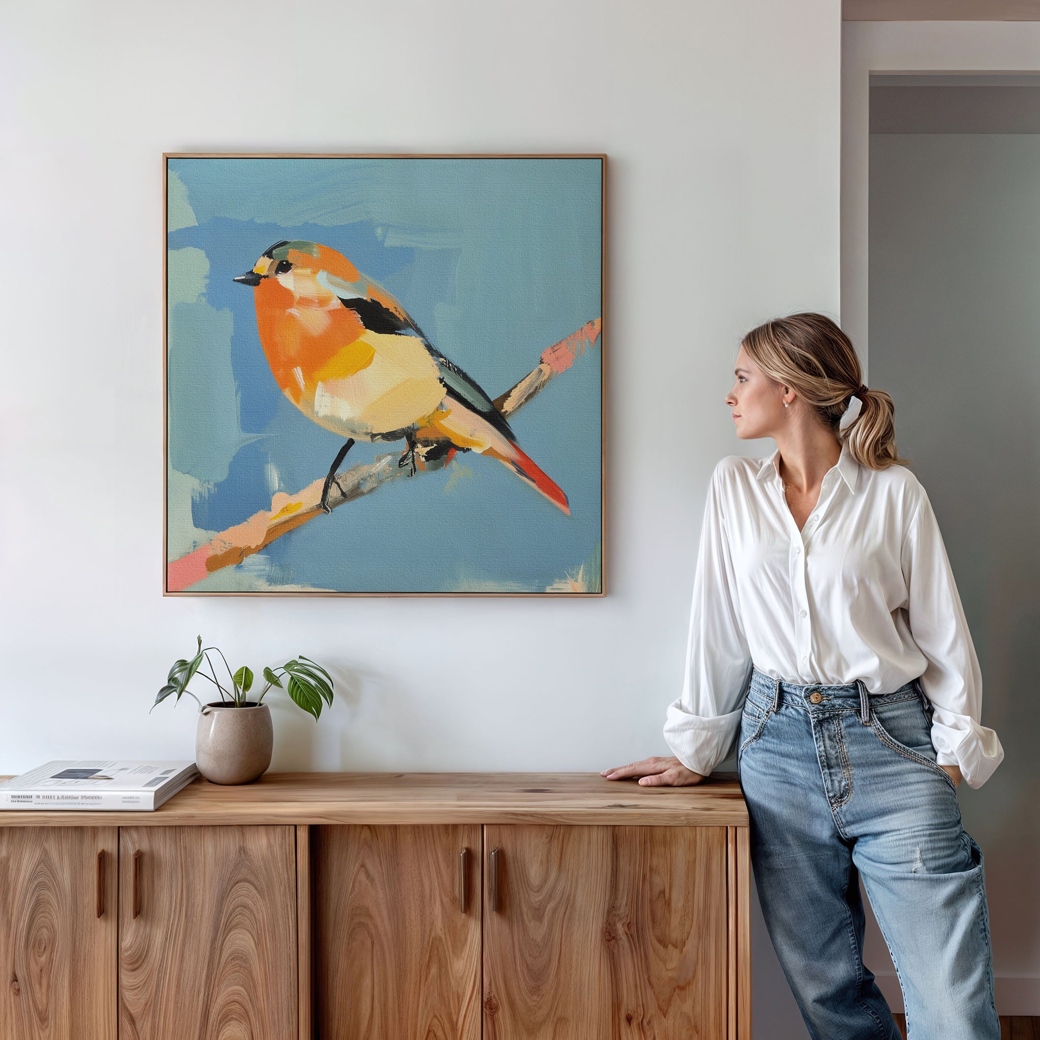A woman in a white blouse and jeans leans against a wooden cabinet, gazing at the Bright Songbird Canvas Art - Square, a colorful bird print on a branch, hanging on the light-colored wall above her.