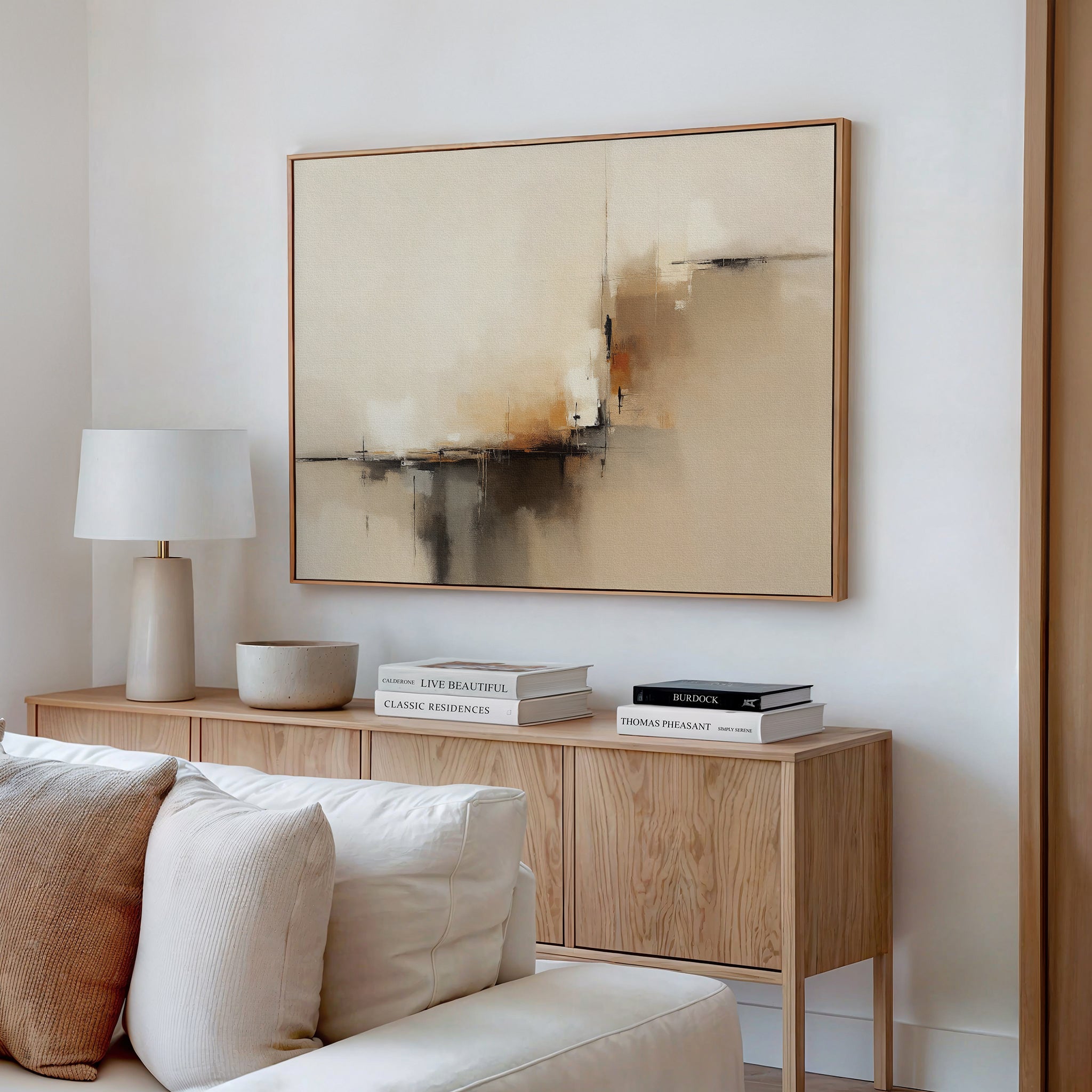 A minimalist living room showcases a wooden sideboard with books, a bowl, and a white lamp. Above hangs the Burnt Sienna Convergence Canvas Art - Horizontal in neutral tones, while a cream-colored sofa is placed in the foreground.