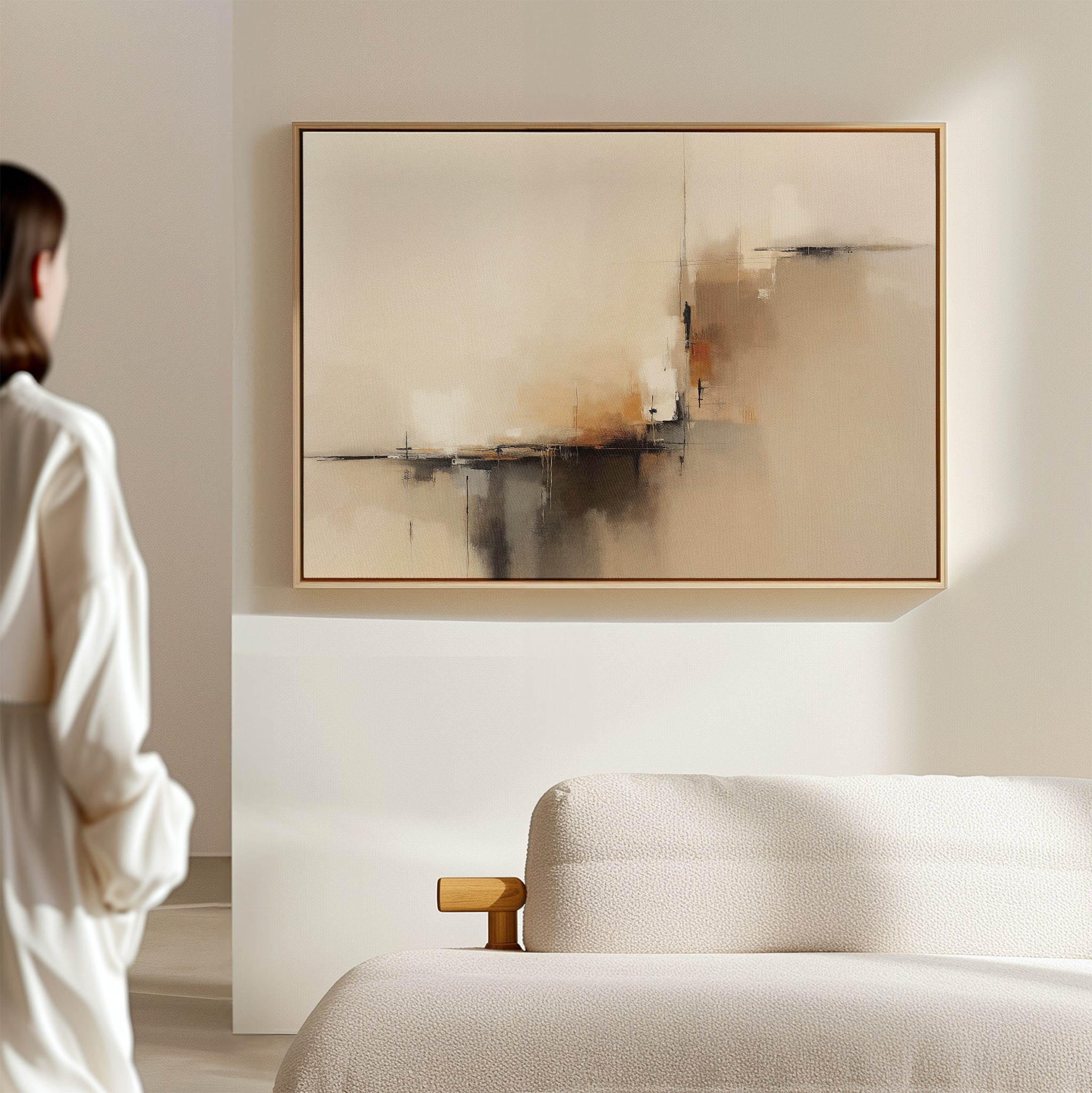 A person in a white robe stands in a sunlit, minimal room, admiring the Burnt Sienna Convergence Canvas Art - Horizontal featuring beige, black, and burnt sienna tones above a light-colored sofa.
