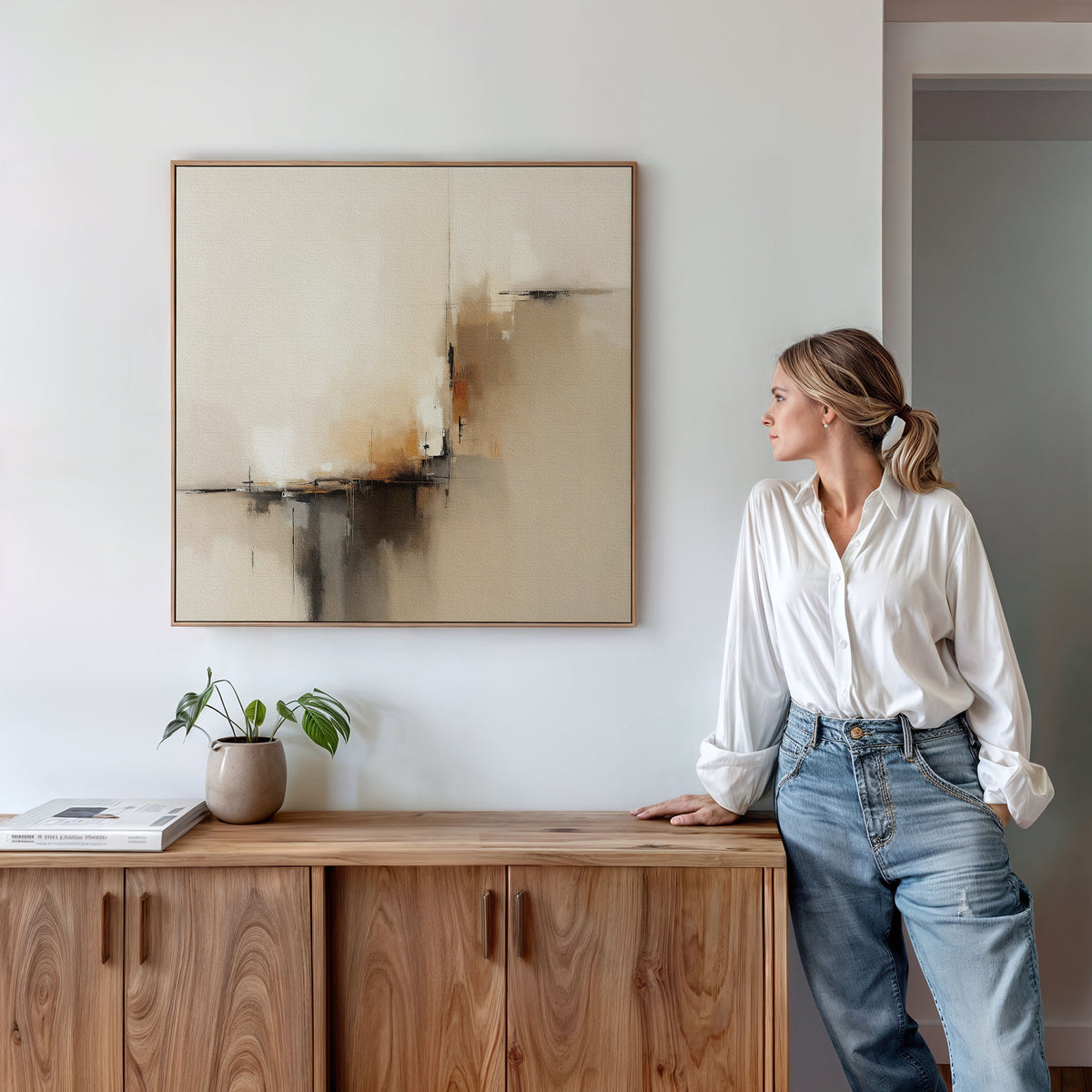 A woman in a white blouse and jeans leans against a wooden cabinet, admiring the Burnt Sienna Convergence Canvas Art - Square. A potted plant and book rest on the cabinet, enhancing the Nordic Park-inspired, neutral-toned decor.