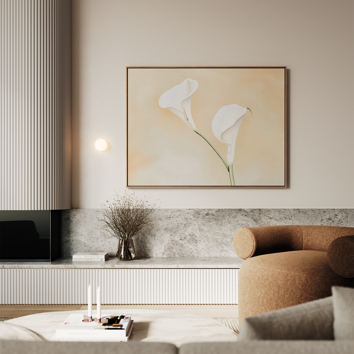 A modern living room with a beige sofa, a marble-topped low cabinet, a glass vase with dried branches, and Calla Serenity Abstract Canvas Art—a large framed painting of two white flowers on a soft peach background.