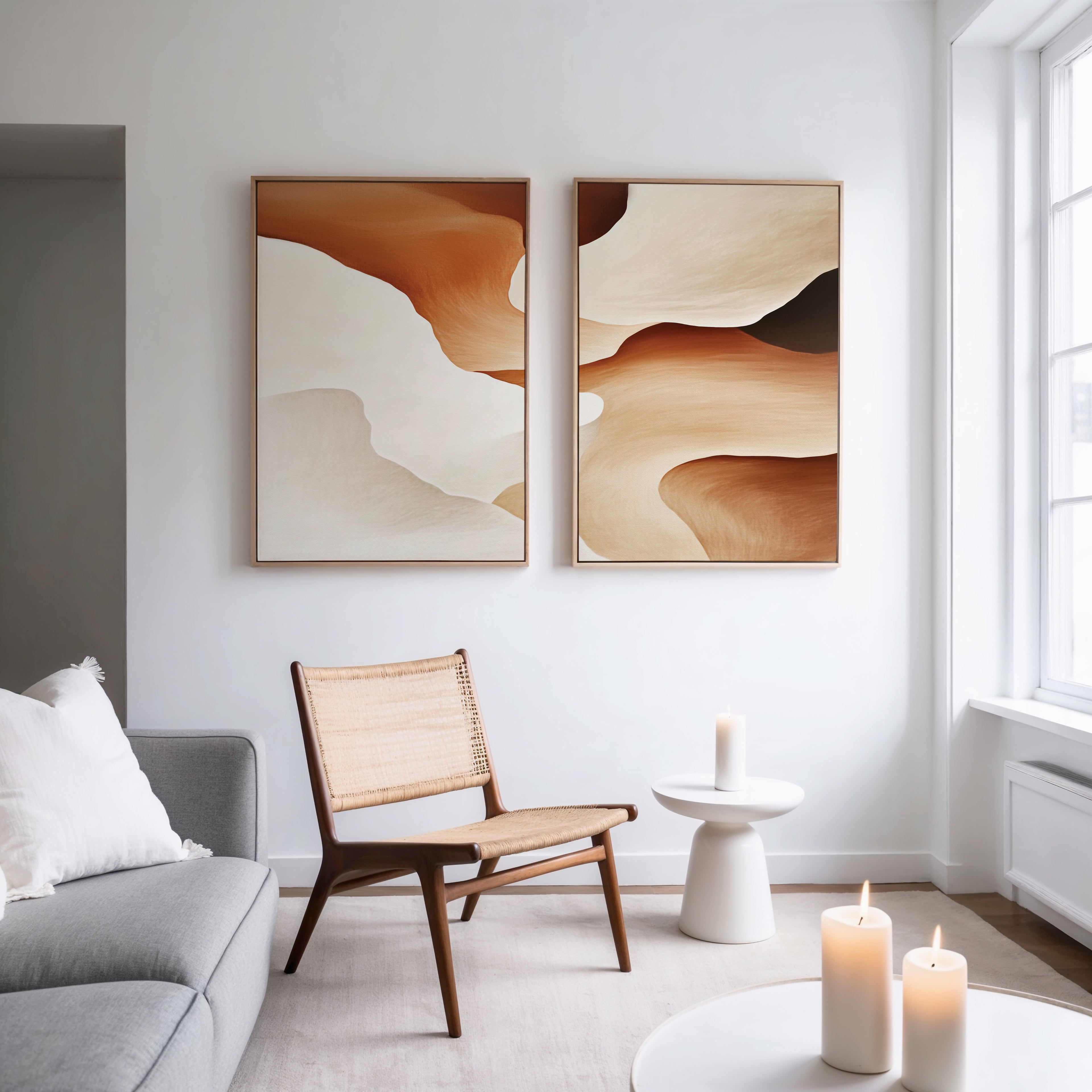 A modern living room features a woven chair, round white side table, lit candles, gray sofa, and the Canyon Curves - Set Of 2 wall art in brown and beige, adding style to the white wall near large windows.