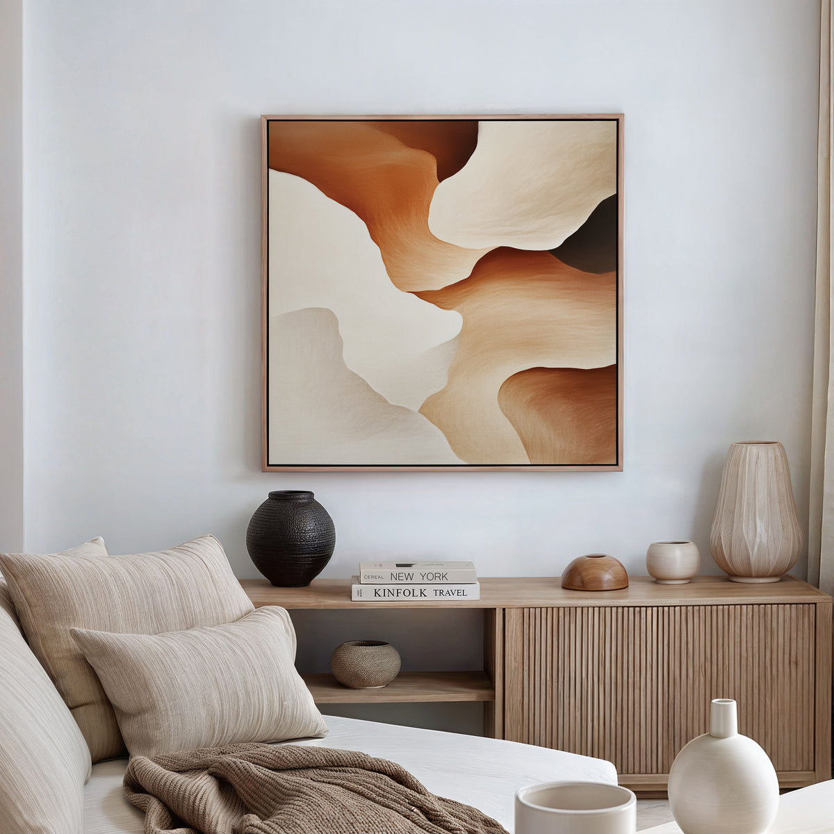 A neutral-toned living room features the Canyon Curves Canvas Art - Square, in beige, brown, and cream above a wooden console with books and decor, next to a beige sofa topped with striped cushions.