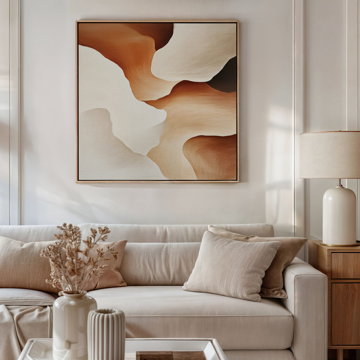 A neutral living room features a beige sofa with matching pillows, a white table lamp on a wooden side table, dried flowers in a vase, and the Canyon Curves Canvas Art - Square—a contemporary abstract in warm earth tones—above the sofa.