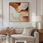 A neutral living room features a beige sofa with matching pillows, a white table lamp on a wooden side table, dried flowers in a vase, and the Canyon Curves Canvas Art - Square—a contemporary abstract in warm earth tones—above the sofa.