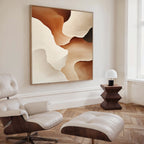 A modern living room features a white leather lounge chair and ottoman, a geometric dark wood side table, a small lamp, and the Canyon Curves Canvas Art - Square—an abstract painting in warm beige and brown tones—to complete the look.