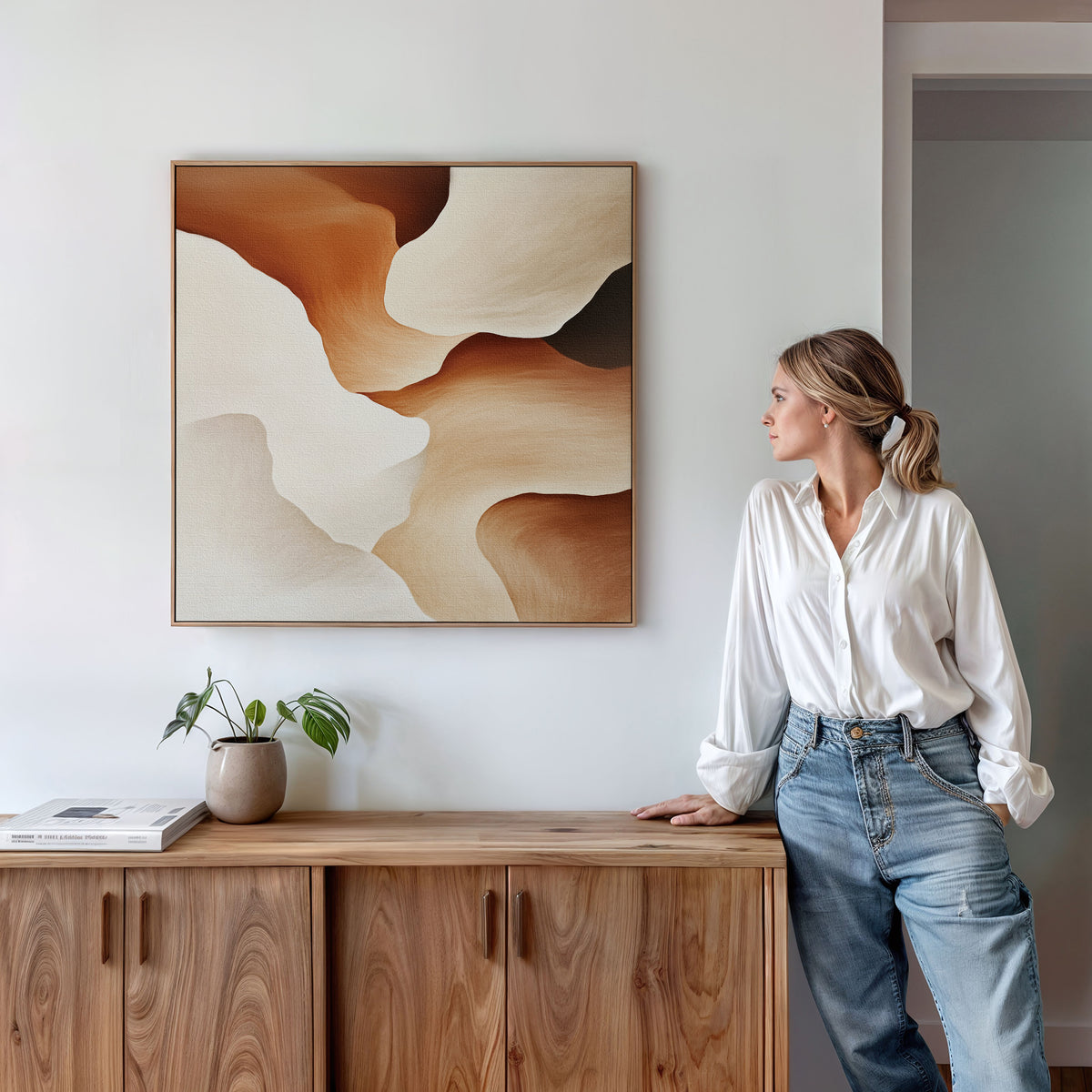 A woman in a white shirt and jeans leans against a wooden sideboard, gazing at the Canyon Curves Canvas Art - Square in brown, beige, and cream. A small plant and books rest on the sideboard beneath the contemporary artwork.