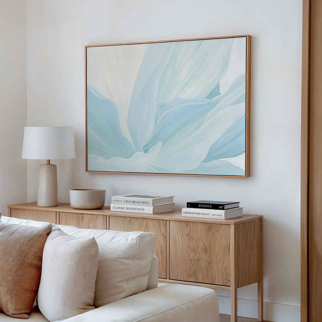 A modern living room features a light wood sideboard, books, a white lamp, beige bowl, and the Icy Petals Abstract Canvas Art in blue and white above. A cream sofa is partially visible, reflecting stylish decor.