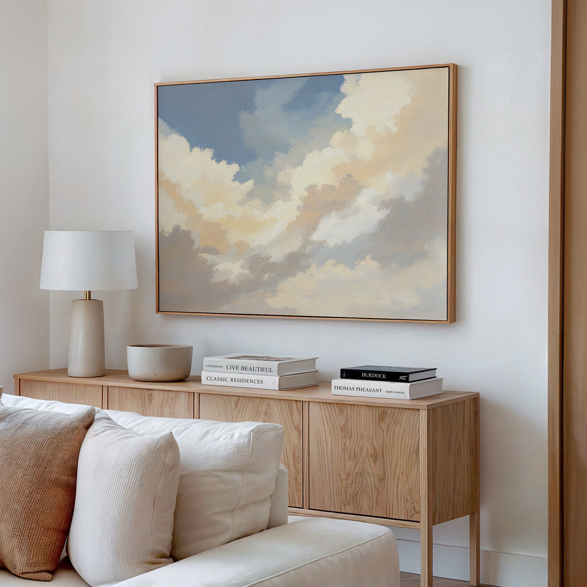 A modern living room with a light wood console table, books, a white lamp, and a bowl features the Celestial Embrace Canvas Art - Horizontal above the console. A cream-colored sofa sits in the foreground.