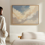 A woman in a white blouse admires the Celestial Embrace Canvas Art - Horizontal, featuring soft clouds and blue sky, hanging above a beige sofa and adding tranquil Nordic vibes to the light-colored, minimalist room.