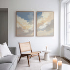 A modern, minimal living room features a light gray sofa, wooden chair, small white side table, and lit candles. Above the chair on a white wall hang two framed pieces from the "Celestial Embrace - Set of 2" collection. Natural light fills the space.