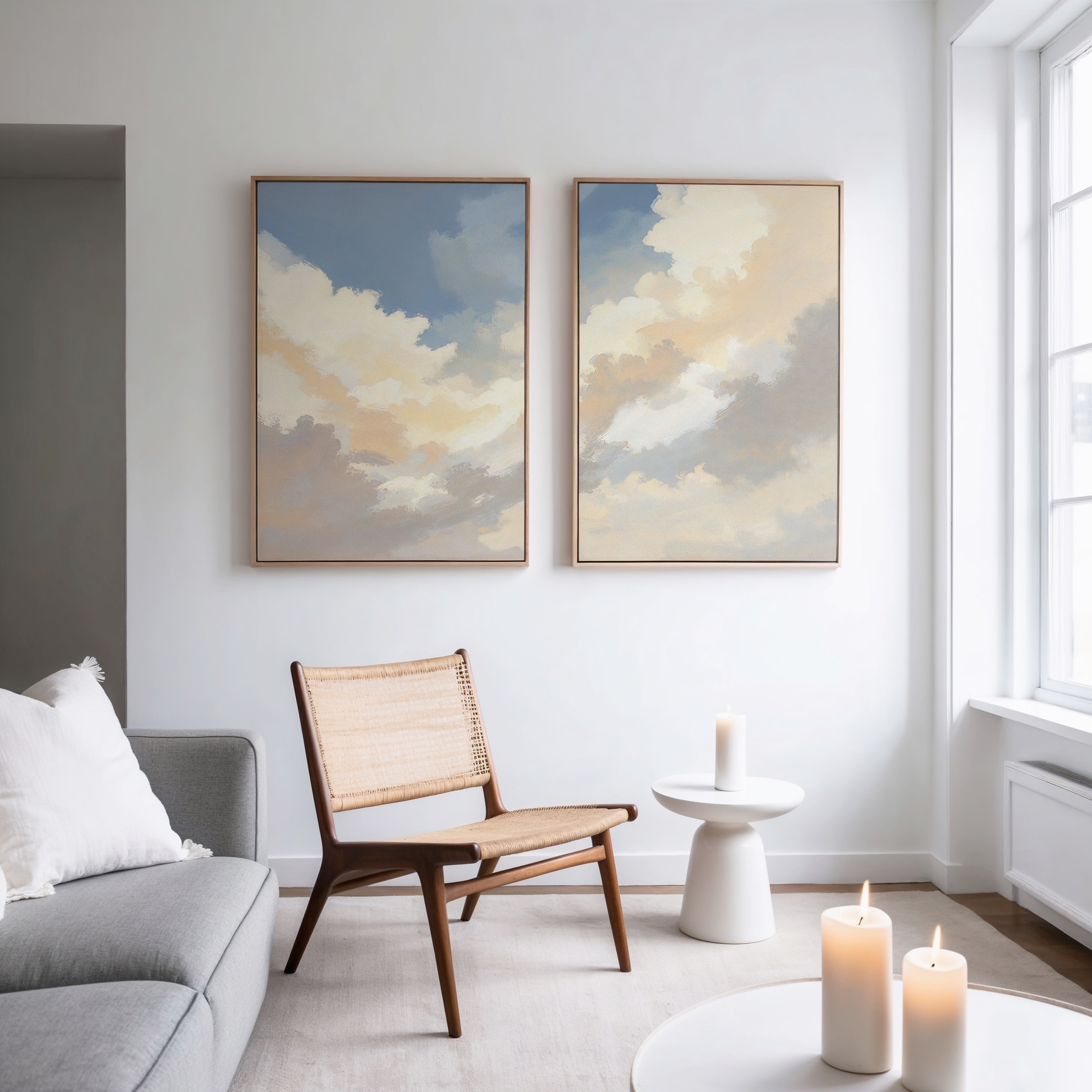 A modern, minimal living room features a light gray sofa, wooden chair, small white side table, and lit candles. Above the chair on a white wall hang two framed pieces from the "Celestial Embrace - Set of 2" collection. Natural light fills the space.