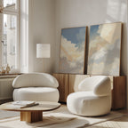 A modern living room with two rounded white armchairs, a wooden coffee table, a floor lamp, and the Celestial Embrace - Set of 2 cloud wall art displayed on a sideboard against a white wall. Sunlight streams through tall windows.