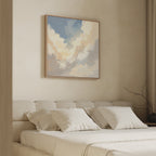 A minimalist bedroom features a beige upholstered bed with white bedding and pillows. Above the bed hangs the Celestial Embrace Canvas Art - Square, depicting clouds in a blue sky. A simple branch decorates the small bedside table.