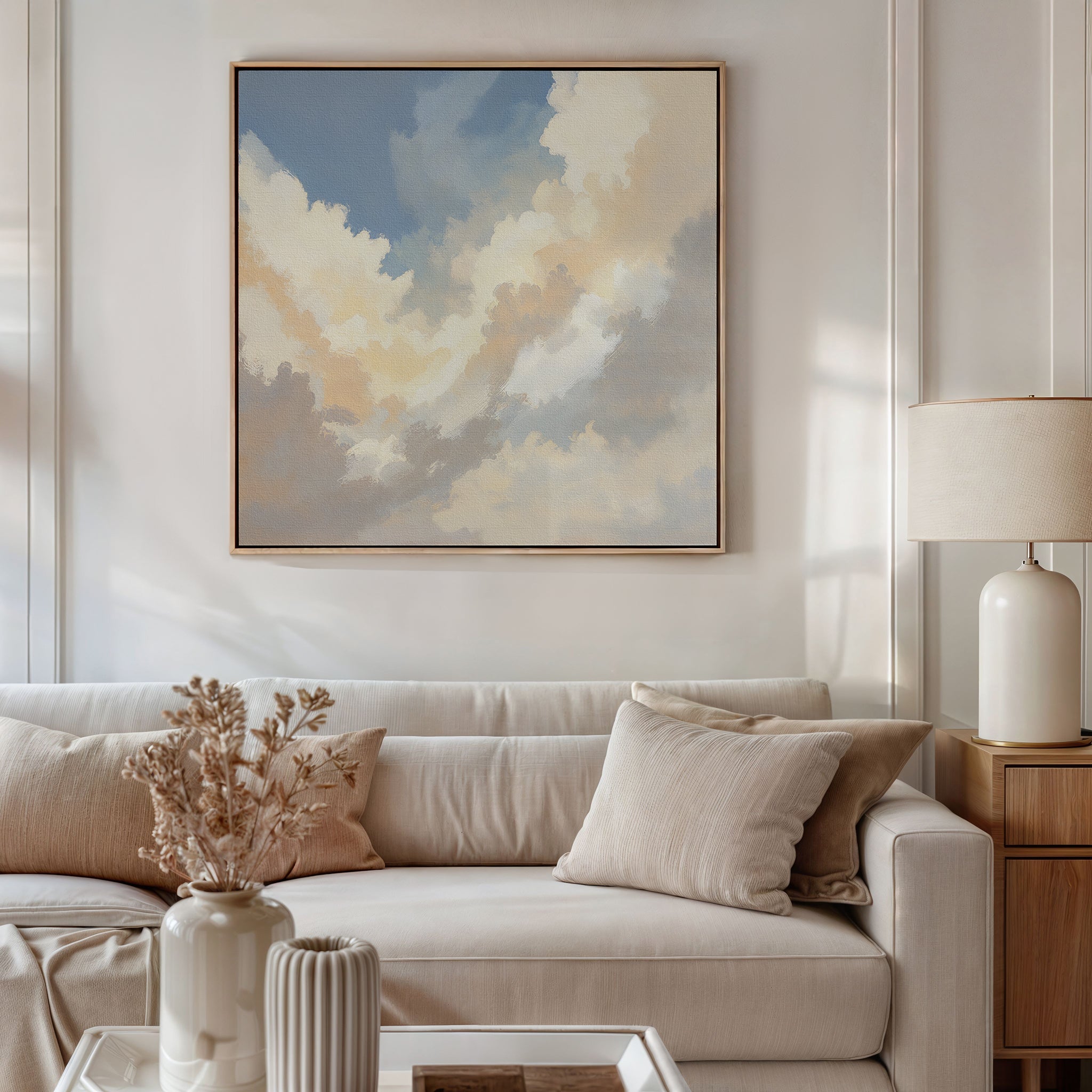 A neutral-toned living room features a beige sofa, cream pillows, light wood side table with lamp, and a vase of dried flowers. Above the sofa hangs the Celestial Embrace Canvas Art - Square, adding celestial charm to the serene decor.