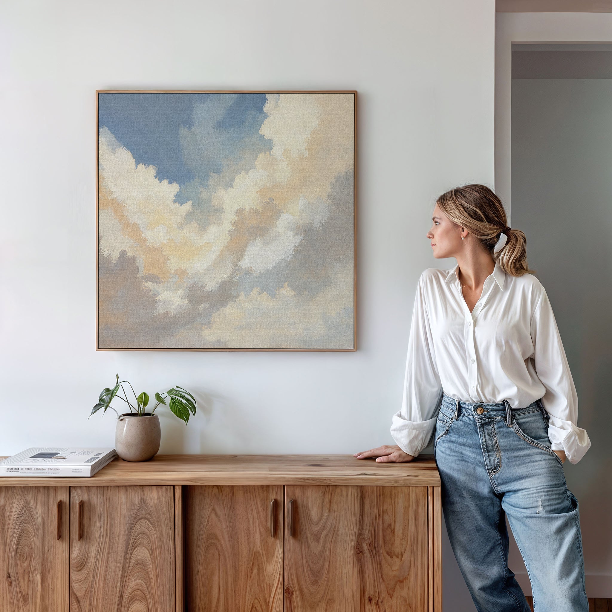 A woman in a white blouse and jeans stands by a wooden cabinet, gazing at the Celestial Embrace Canvas Art - Square above her. A potted plant and book accentuate the calm, minimalistic style of this serene room.