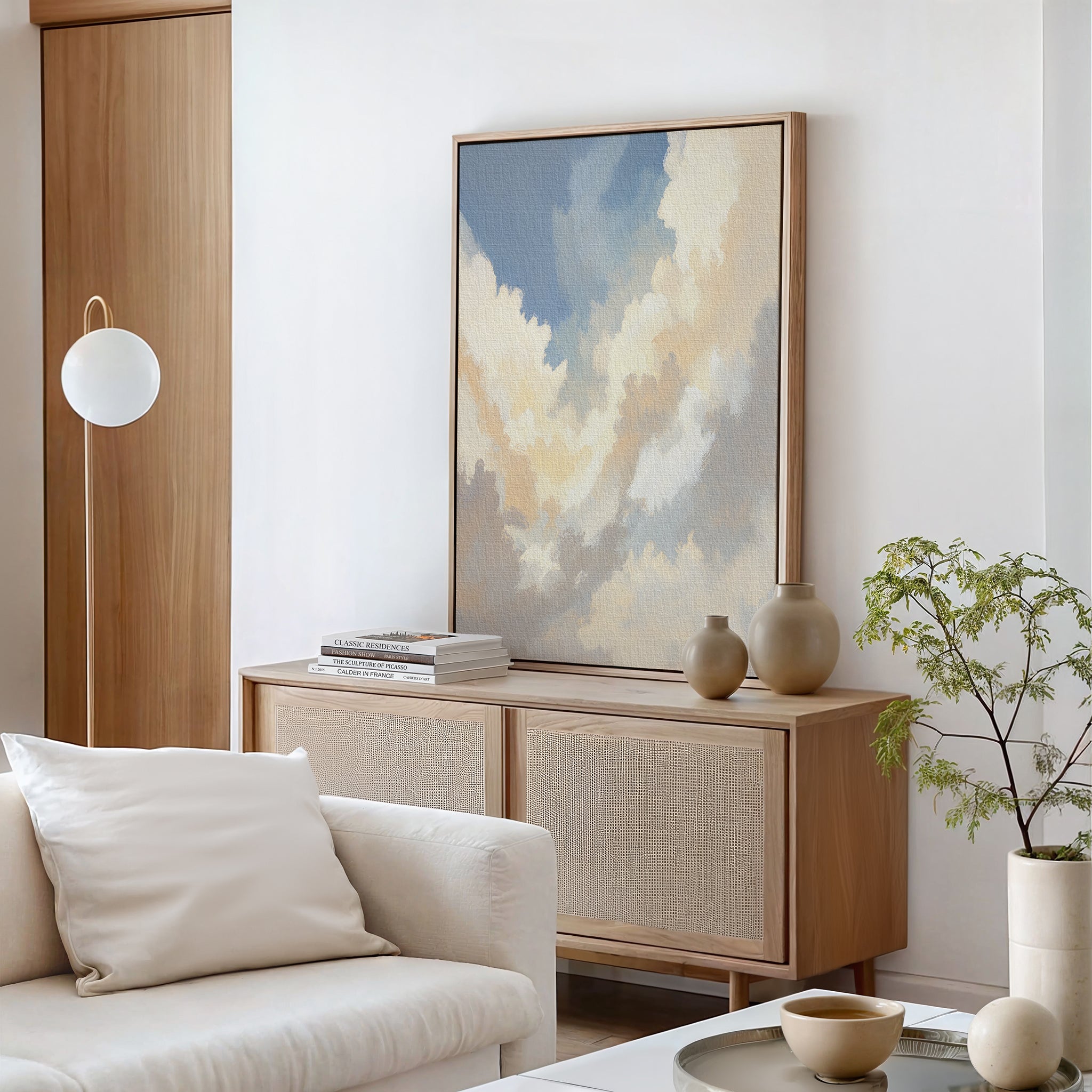 A modern living room features a light wood cabinet, decorative vases, stacked books, the Celestial Embrace Canvas Art - Vertical as serene wall art, a white cushioned sofa, a round hanging lamp, and a small potted plant.