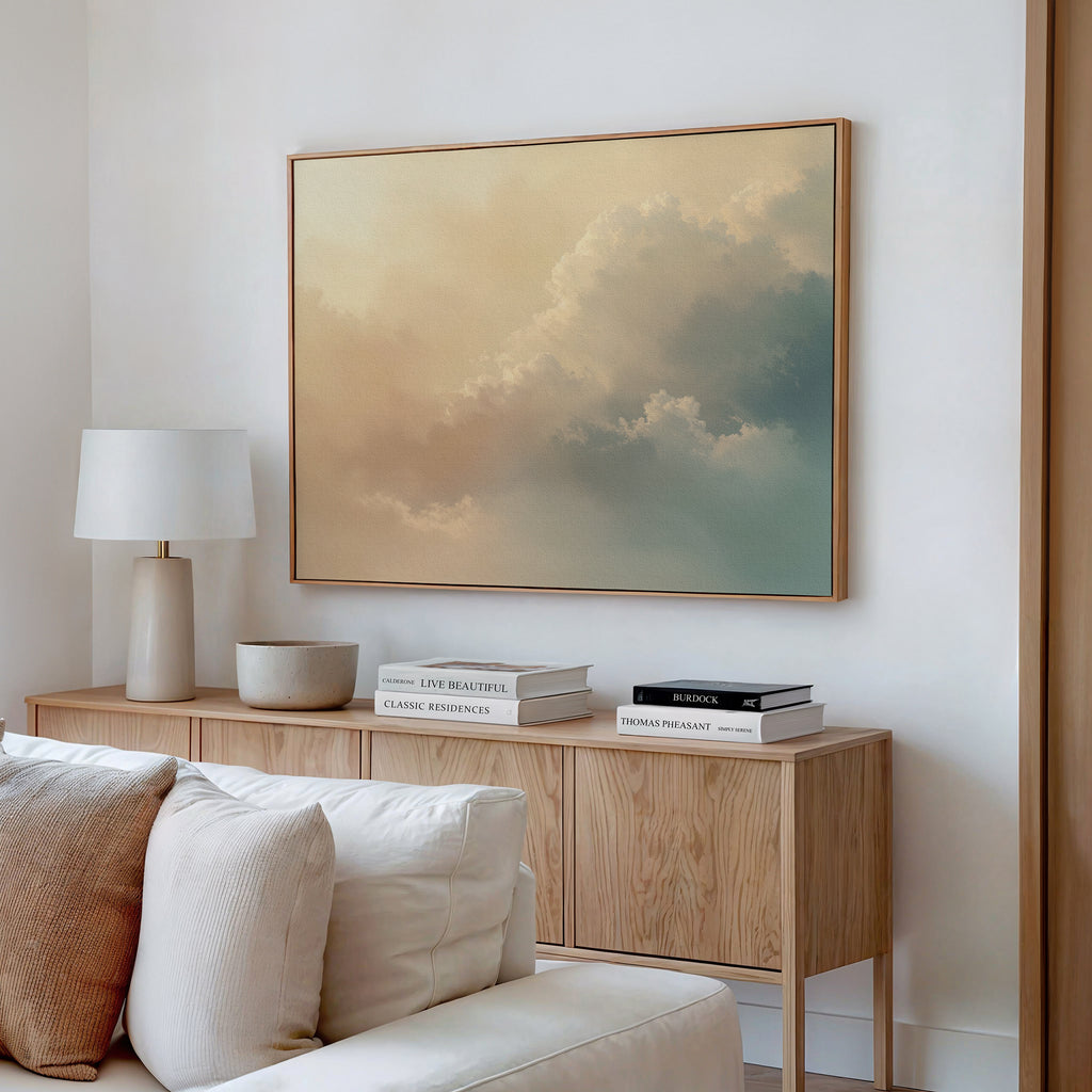 A minimalist living room features a wooden sideboard, white lamp, decorative bowl, stacked books, and the Celestial Veil Canvas Art - Horizontal with soft pastel clouds above. A white couch is partially visible in the foreground.