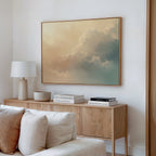 A minimalist living room features a wooden sideboard, white lamp, decorative bowl, stacked books, and the Celestial Veil Canvas Art - Horizontal with soft pastel clouds above. A white couch is partially visible in the foreground.