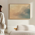 A person in white stands in a minimal, sunlit room featuring champagne light, a beige sofa, and the Celestial Veil Canvas Art - Horizontal with soft pastel clouds hanging on the wall.