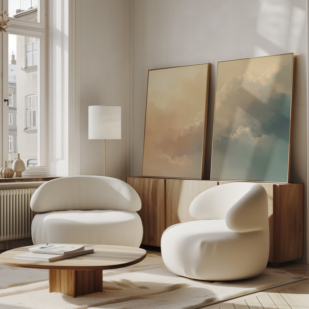 A minimalistic living room features two modern white curved armchairs, a low wooden table, and a wooden cabinet topped with Celestial Veil - Set of 2 prints in champagne tones, positioned by a tall window flooding the space with natural light.
