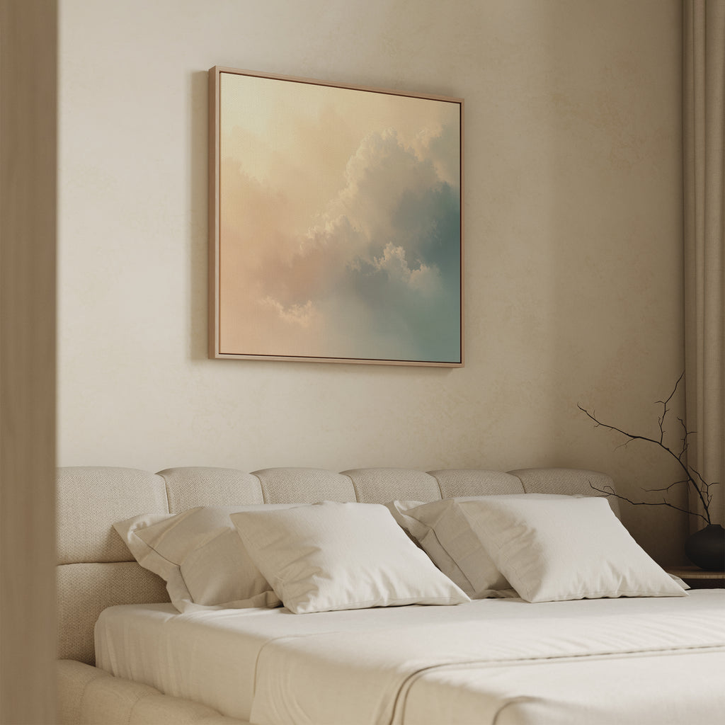 A minimalist bedroom features a beige upholstered bed with white linens. Above the bed hangs the Celestial Veil Canvas Art - Square, showcasing soft, pastel clouds. A simple branch accents the nightstand.