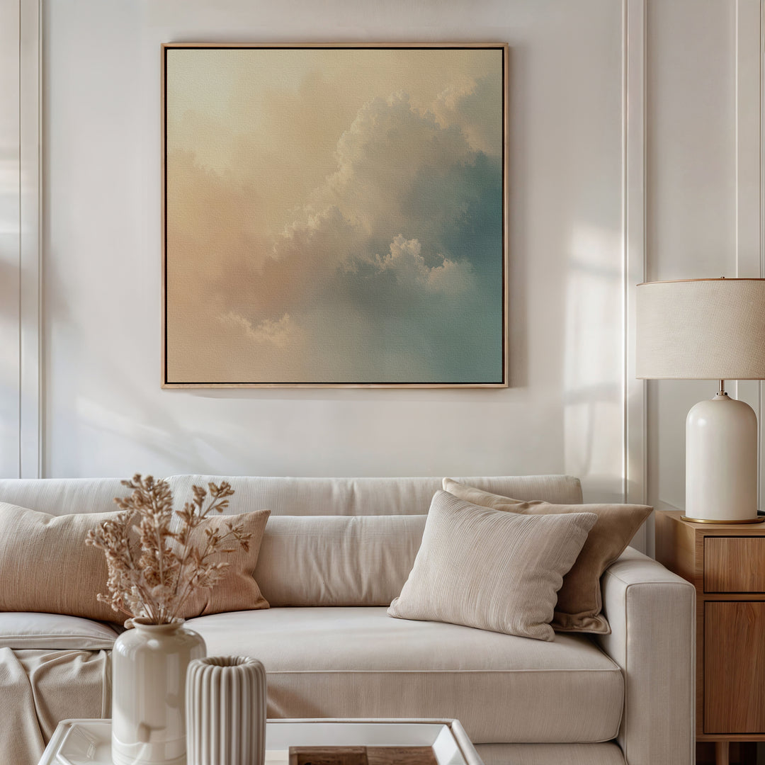 A modern living room features a beige sofa, neutral cushions, a white table lamp, and dried flowers. Above the sofa hangs the Celestial Veil Canvas Art - Square, with pastel clouds that add tranquility to the minimalist decor.
