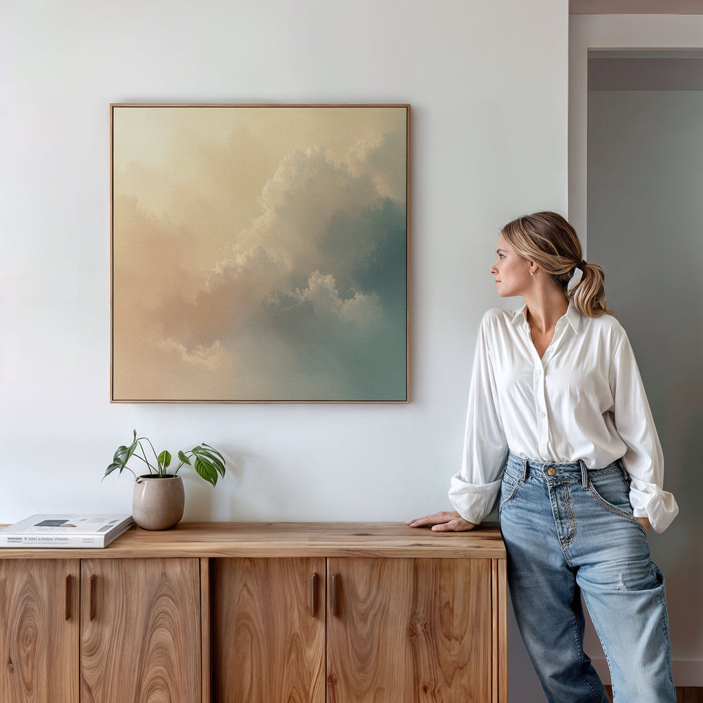 A woman in a white blouse and jeans leans against a wooden cabinet, gazing at the Celestial Veil Canvas Art - Square, which depicts soft clouds in champagne light. A potted plant and books sit on the cabinet under the serene artwork.