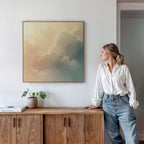 A woman in a white blouse and jeans leans against a wooden cabinet, gazing at the Celestial Veil Canvas Art - Square, which depicts soft clouds in champagne light. A potted plant and books sit on the cabinet under the serene artwork.