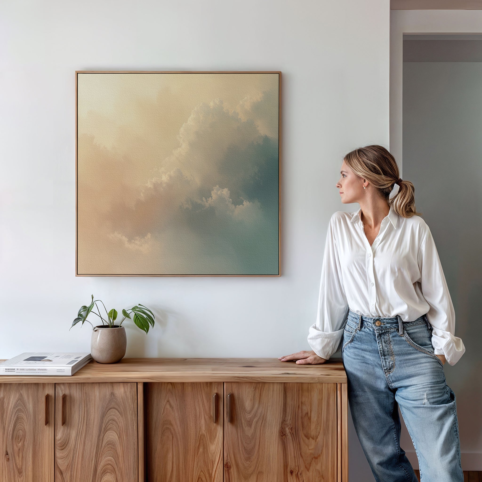 A woman in a white blouse and jeans leans against a wooden cabinet, gazing at the Celestial Veil Canvas Art - Square, which depicts soft clouds in champagne light. A potted plant and books sit on the cabinet under the serene artwork.
